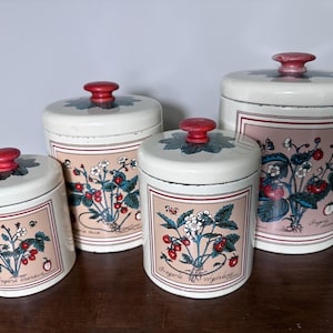 May include: Set of four white ceramic canisters with red knobs and strawberry plant illustrations. Each canister has a pink rectangular label with the botanical name "Fragaria virginiana". The lids have a green leaf design.