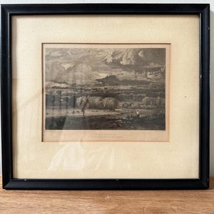 May include: Framed black and white landscape print. The artwork depicts a river scene with trees, figures, and a cloudy sky. The print is set within a cream-colored mat and a black wooden frame.