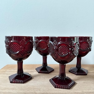 May include: Four ruby red goblets with ornate, textured designs. Each goblet features a round bowl, a slender stem, and a hexagonal base. The goblets are arranged on a light brown wooden surface.