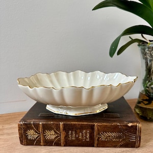 May include: An off-white ceramic bowl with a scalloped edge and a gold-painted rim. The bowl features a fluted, shell-like design and rests on a small, oval-shaped pedestal base with a matching gold-painted trim along the bottom edge.