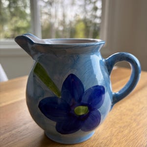 May include: A small, round, blue ceramic pitcher with a handle and spout. The pitcher is decorated with a large, dark blue flower and green leaves. The background is a light blue wash.