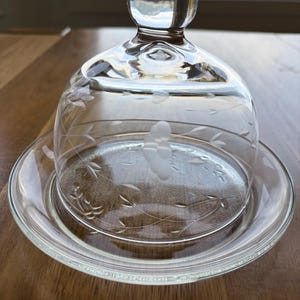 May include: Clear glass butter dish with a domed lid and etched floral design. The dish includes a round base and a knob on top of the lid. The glass is transparent, allowing the contents to be visible. The dish is designed for serving butter.