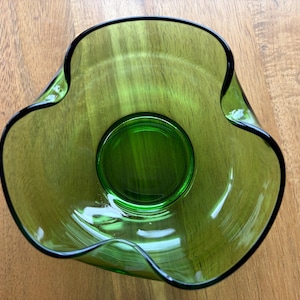 Op de afbeelding: Een doorschijnende groene glazen kom met een brede, golvende en kronkelende rand. De kom heeft een gladde, ronde bodem en een diepe binnenkant, met een levendige smaragdgroene tint door het glas heen en een donkere rand.