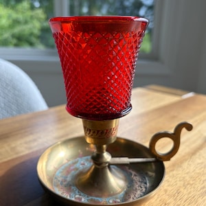 Può includere: Un portacandele in ottone caratterizzato da una base circolare con un motivo inciso decorativo e un manico laterale. Un vasetto votivo in vetro rosso vivace con una superficie testurizzata a rombi poggia saldamente al centro del supporto.