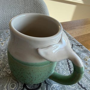 May include: A ceramic mug with a distinctive design. The mug has a white interior and a light green exterior with a speckled pattern. The handle is shaped like a whale's tail, adding a whimsical touch. Suitable for hot or cold drinks.