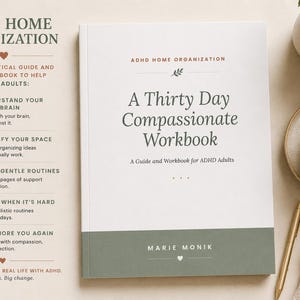 Könnte beinhalten: Ein weißes Arbeitsbuch mit dem Titel "A Thirty Day Compassionate Workbook" mit dem Text "ADHD Home Organization". Das Buch liegt auf einer hellen Oberfläche mit einer Tasse Kaffee, einem Stift und einer Pflanze.
