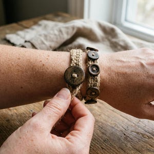 May include: Two bracelets made of woven tan jute twine. One bracelet features a single large, dark brown circular wooden button as a closure. The second bracelet is adorned with a row of three smaller, dark brown circular wooden buttons.