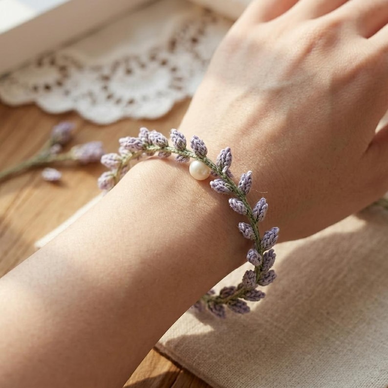 May include: A crochet bracelet featuring a vine-like design with small, light purple thread-stitched leaves attached to a thin green stem, accented by a single, round, white pearl bead positioned along the length of the delicate floral band.