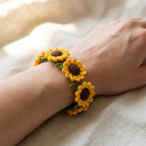 May include: A crochet bracelet featuring a series of connected sunflower motifs. Each flower has a dark brown center surrounded by bright yellow petals and small green leaves attached between the blooms, creating a continuous floral chain design.