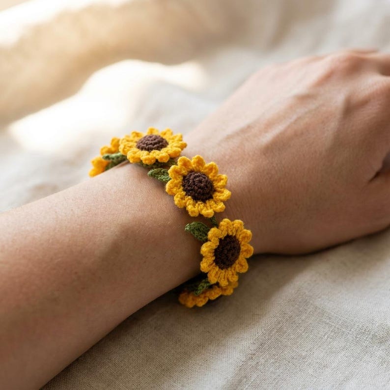 May include: A crochet bracelet featuring a series of connected sunflower motifs. Each flower has a dark brown center surrounded by bright yellow petals and small green leaves attached between the blooms, creating a continuous floral chain design.