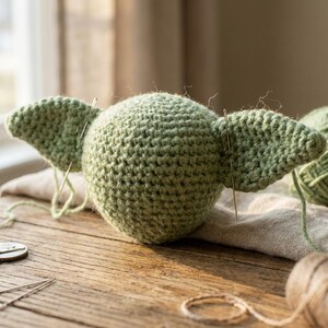 May include: A light green crocheted figure with large ears, possibly a character from a popular science fiction series. The figure is in the process of being assembled, with needles inserted into the ears. The project is on a wooden surface.