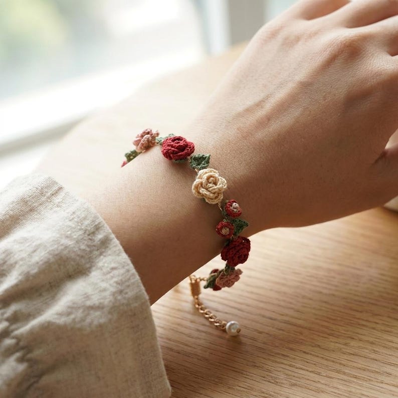 May include: A bracelet featuring small, crocheted floral motifs in shades of red, pink, and cream, interspersed with tiny green crocheted leaves. The bracelet includes a gold-toned metal chain extension with a small white bead at the end.