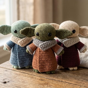 May include: Three handmade crochet dolls in the style of a popular character. Each doll has large ears, black eyes, and a robe. The dolls are in shades of blue, orange, and burgundy. The dolls are displayed on a wooden surface.