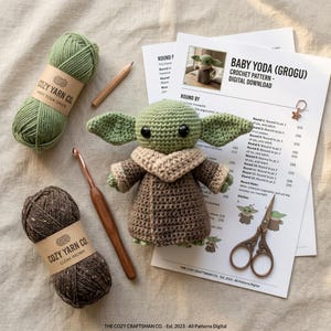 May include: A crocheted Baby Yoda (Grogu) doll with large ears, brown robe, and tan collar sits next to green and brown yarn, a crochet hook, scissors, and a pattern titled "Baby Yoda (Grogu) Crochet Pattern - Digital Download."