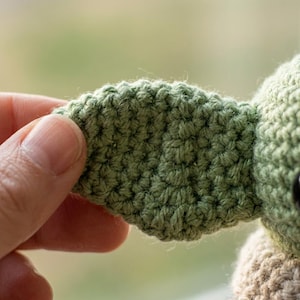 May include: Close-up of a crocheted green ear of a doll. The ear is held by a hand, revealing the detailed texture of the yarn. The doll's head and body are also visible, with a tan body and a black eye.