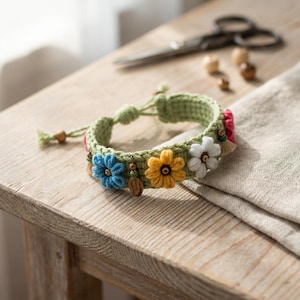 May include: A sage green crocheted bracelet featuring three-dimensional embroidered flowers in blue, yellow, and white. The band is accented with small wooden beads and dangling charms, finished with a sliding knot closure for an adjustable fit.