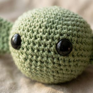 May include: Close-up of a crocheted green ball with two large, glossy black eyes. The handmade item is likely a toy or decorative piece, showcasing the texture of the yarn and the detailed craftsmanship.