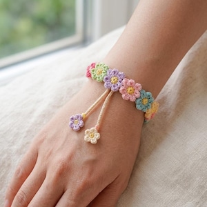 May include: A crochet bracelet featuring a series of small, multi-colored flowers in shades of pink, green, purple, blue, and yellow. The bracelet includes two dangling flower charms attached to thin, light-colored braided cords for adjustment.