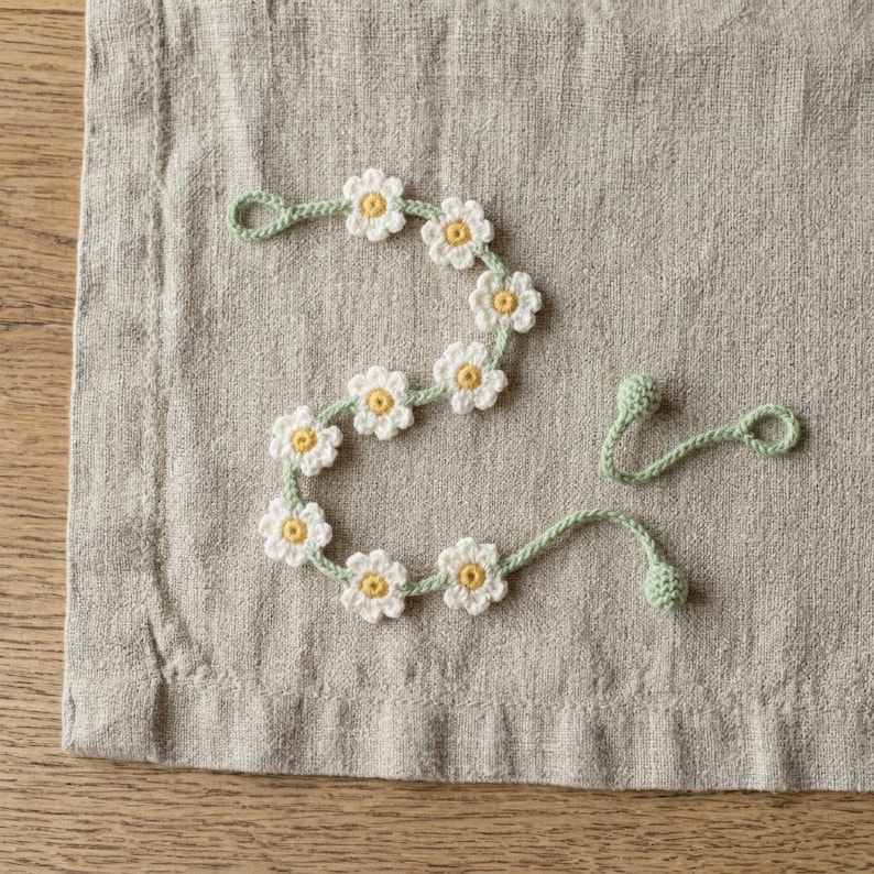 May include: A crochet garland featuring a series of white flowers with yellow centers connected by a light green vine. The vine ends in small green loops and spherical knots, creating a continuous floral strand with a soft, textured appearance.