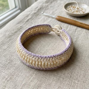 May include: A circular crochet bracelet featuring cream-colored yarn and small, translucent beads woven throughout. The edges are trimmed in a soft lavender yarn, and the piece is secured with a small, round, off-white button closure.