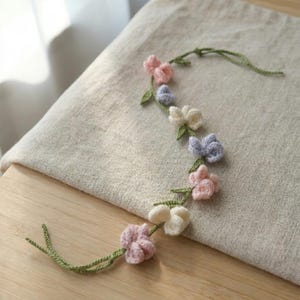 May include: A crocheted garland featuring a thin green vine with small, soft-textured flowers in shades of light pink, pale lavender, and cream. The floral vine has long, slender green stems extending from both ends, creating a delicate, flexible decorative strand.