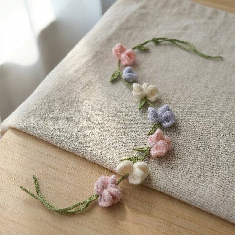 May include: A crocheted garland featuring a thin green vine with small, soft-textured flowers in shades of light pink, pale lavender, and cream. The floral vine has long, slender green stems extending from both ends, creating a delicate, flexible decorative strand.