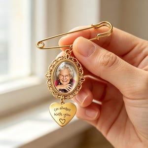 Puede incluir: Un broche de imperdible dorado con un marco ovalado de borde filigranado que contiene la fotografía de una persona mayor con gafas. Un pequeño dije dorado en forma de corazón cuelga debajo con el texto I'm always with you.