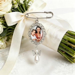 Puede incluir: Un broche de metal plateado con un marco ovalado y un borde de filigrana ornamentado. El marco contiene una fotografía de una mujer y un niño. Un pequeño dije colgante con forma de ángel y un cuerpo de perla blanca en forma de lágrima cuelga debajo.