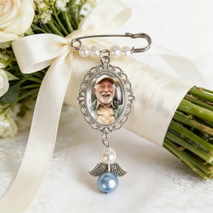 May include: A silver-toned brooch pin featuring an oval frame containing a photograph of a smiling person wearing a cap and collared shirt. The pin is adorned with white faux pearls and a dangling charm with a white pearl and a light blue bead.
