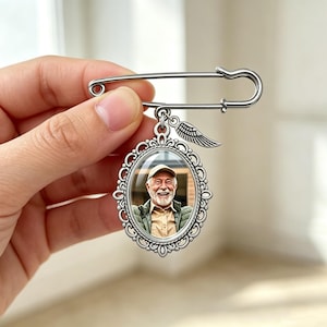 Puede incluir: Un broche de imperdible plateado con un colgante ovalado de marco de filigrana ornamentado. El colgante contiene la fotografía de una persona mayor sonriente con camisa beige y gorra. Un pequeño dije de ala plateada cuelga de la parte superior.