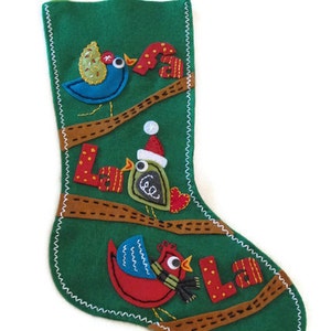 Puede incluir: Calcetín de Navidad de fieltro verde con un diseño cosido de tres pájaros, dos con acentos rojos y amarillos y uno azul, y la palabra "La La" en letras de fieltro amarillo.