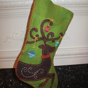 May include: A green felt stocking with a brown reindeer applique and colorful felt ornaments. The reindeer has a red and white collar and a brown swirl design on its back.