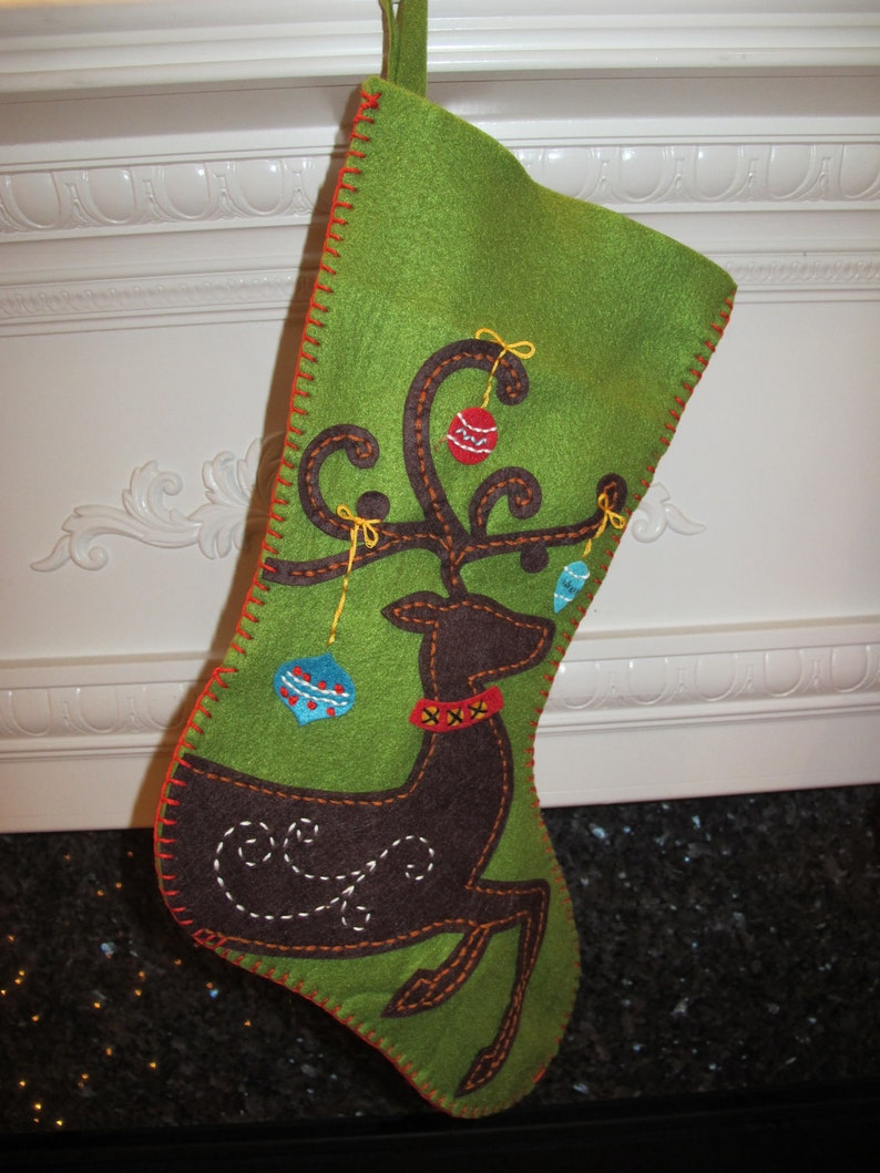 May include: A green felt stocking with a brown reindeer applique and colorful felt ornaments. The reindeer has a red and white collar and a brown swirl design on its back.