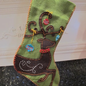May include: Green felt Christmas stocking with a brown felt reindeer and red, blue, and white ornaments. The stocking has a red and orange stitched border.