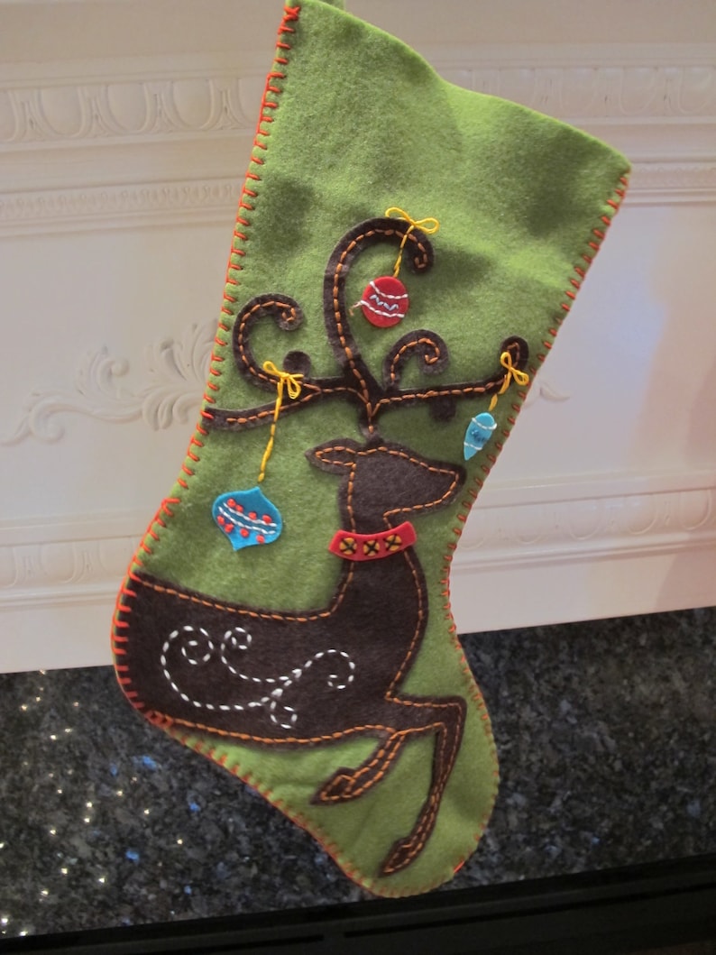 May include: Green felt Christmas stocking with a brown felt reindeer and red, blue, and white ornaments. The stocking has a red and orange stitched border.