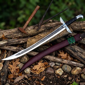 Könnte beinhalten: Ein silbernes und schwarzes Schwert mit gebogener Klinge, das auf einem Stapel Holzstämme liegt. Das Schwert hat einen dekorativen Knauf und wird von einer weinroten Lederscheide begleitet. Ein Hammer und eine Zange sind ebenfalls sichtbar.