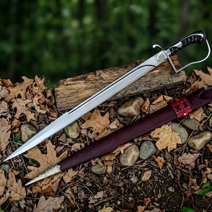 May include: A silver sword with a black and silver handle, resting on a wooden log. The sword has a curved blade and a silver handguard. A burgundy leather sheath with red stitching lies beside it, set against a backdrop of autumn leaves and forest.