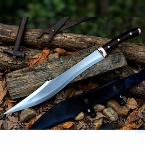 May include: A large, silver blade knife with a dark wooden handle and copper accents. The knife rests on a pile of wood with a black leather sheath. A hammer and pliers are visible in the background.