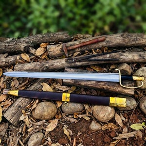Könnte beinhalten: Ein langes, silbernes Schwert mit schwarzem Griff und goldenen Akzenten, das auf einem Bett aus Blättern und Steinen liegt. Eine braune Lederscheide mit goldenen Zierleisten liegt darunter. Ein Hammer und eine Zange sind im Hintergrund zu sehen.