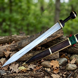 Könnte beinhalten: Ein römisches Gladius-Schwert mit polierter Stahlklinge und dunklem Holzgriff. Der Knauf hat goldfarbene Akzente. Das Schwert liegt auf einem Bett aus Baumstämmen und Herbstblättern, mit seiner Holzscheide in der Nähe. Es ist eine historische Replik.