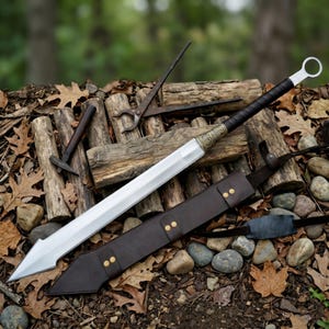 May include: A large, silver sword with a black handle and a decorative gold-coloured hilt, resting on a bed of logs and rocks. A brown leather sheath with gold-coloured rivets accompanies the sword. Additional tools, including a hammer and tongs, are also present.
