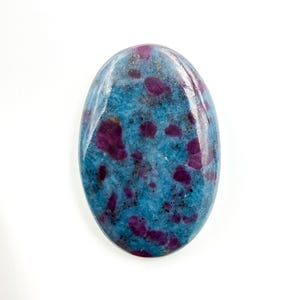 Puede incluir: Una piedra preciosa pulida de forma ovalada con un color base azul vibrante y dispersas inclusiones irregulares de color magenta intenso y morado oscuro por toda la superficie. La piedra tiene un acabado liso y brillante en forma cabujón.