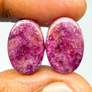 May include: Two oval-shaped cabochons featuring a vibrant magenta and deep pink color palette with a natural crystalline texture. The stones display a polished, smooth surface with intricate internal patterns of white and dark pink mineral inclusions.