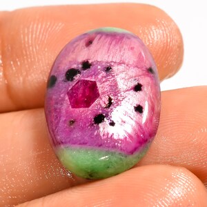 Op de afbeelding: Een ovaalvormige cabochon edelsteen met een levendig magenta en roze centrum met een opvallende zeshoekige dieprode insluiting, geaccentueerd door verspreide zwarte stippen en een effen lichtgroene basis aan de onderkant, glad gepolijst.