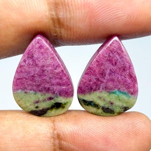 May include: Two pear-shaped cabochons featuring a vibrant magenta-pink upper section and a pale green lower section with dark black inclusions. These polished gemstones display a distinct color-blocked pattern with a smooth, rounded surface.