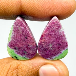 May include: Two pear-shaped cabochons featuring a vibrant combination of deep magenta-pink ruby and bright lime-green zoisite. Each polished gemstone displays a smooth, rounded surface with natural color variations and small black mineral inclusions.
