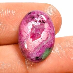 Può includere: Una gemma cabochon di forma ovale caratterizzata da una vibrante tavolozza di colori magenta e rosa intenso, con texture cristalline visibili, inclusioni interne e una piccola macchia di materiale minerale verde vicino al bordo inferiore.