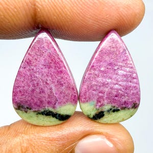 Op de afbeelding: Twee peervormige cabochons met een levendig magenta-roze bovengedeelte met een kristallijne textuur, overgaand in een licht limoengroene basis, gescheiden door een duidelijke, onregelmatige horizontale band van zwarte mineraalinsluitingen.