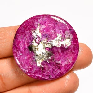 May include: A circular, polished gemstone cabochon featuring a vibrant magenta and deep pink crystalline texture. The center of the stone displays a prominent, irregular patch of metallic silver-colored inclusions that contrast with the surrounding pink.
