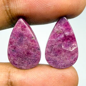 May include: Two pear-shaped, cabochon-cut gemstones with a deep magenta and purple hue. The stones feature a natural, textured surface with visible crystalline inclusions and a polished finish, displaying variations in color intensity throughout.